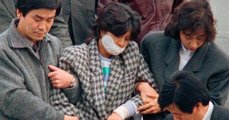 Hatóságiak kísérik Kim Hjon Huit őrizet alatt. A szájába helyezett eszköz célja, hogy megakadályozza, hogy a nő leharapja saját nyelvét. (kép forrása: 2oceansvie.com)