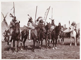 Lóháton ülő amerikai őslakosok 1905 körül. A háttérben látható épületek arra engednek következtetni, hogy a fotó egy vadnyugati előadáson készült.
